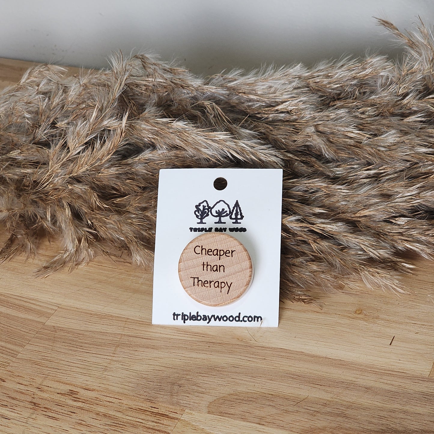 A wooden wine stopper with the phrase 'Cheaper than Therapy' engraved on it, displayed on a card, alongside a rubber version, with a furry item in the background.