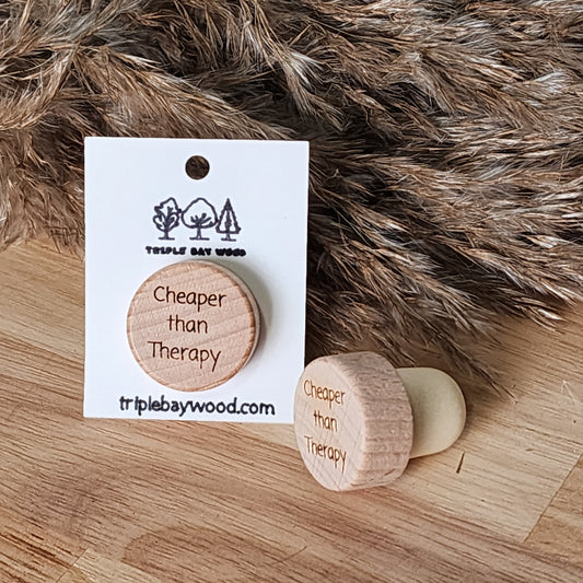 A wooden wine stopper with the phrase 'Cheaper than Therapy' engraved on it, displayed on a card, alongside a rubber version, with a furry item in the background.