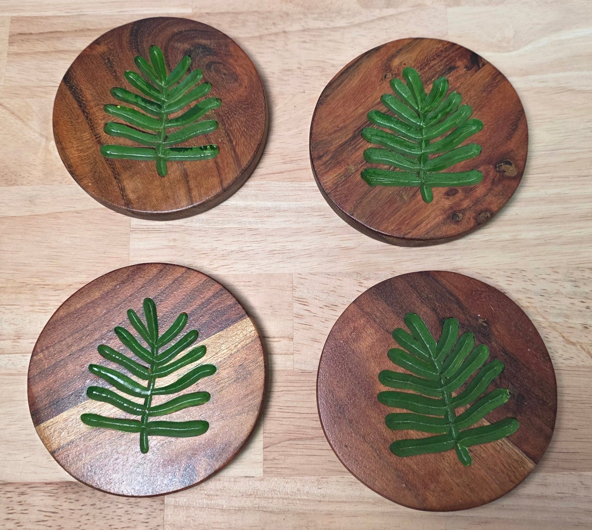 Four wooden coasters with green leaf patterns on a light wood surface.